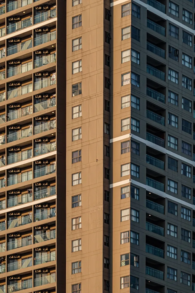 A dense wall of apartment balconies in low golden light, three towers overlapping: one warm and cluttered with air conditioners and laundry, one a flat tan grid of windows, one dark grey with glass balconies