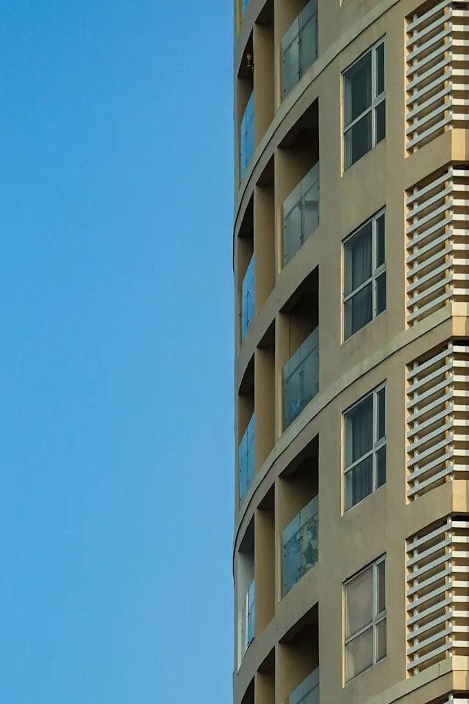 A curved residential tower, half the frame empty blue sky, the other half tan concrete balconies, glass balustrades and white louvre fins repeating up the facade