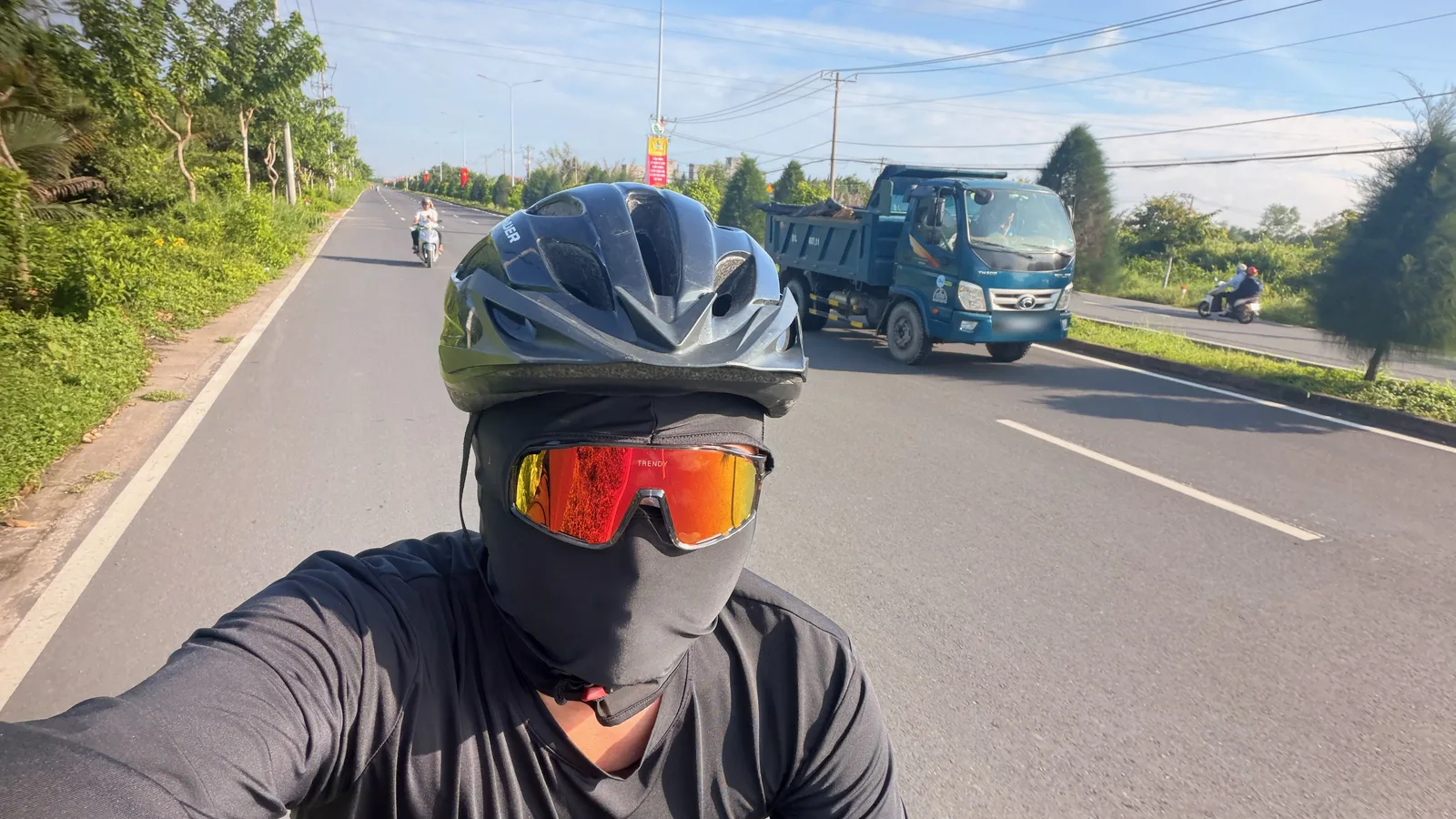 Me in a helmet, balaclava and mirrored sunglasses on the open road to Vũng Tàu, with a blue tipper truck passing on the far side and green verges either way.