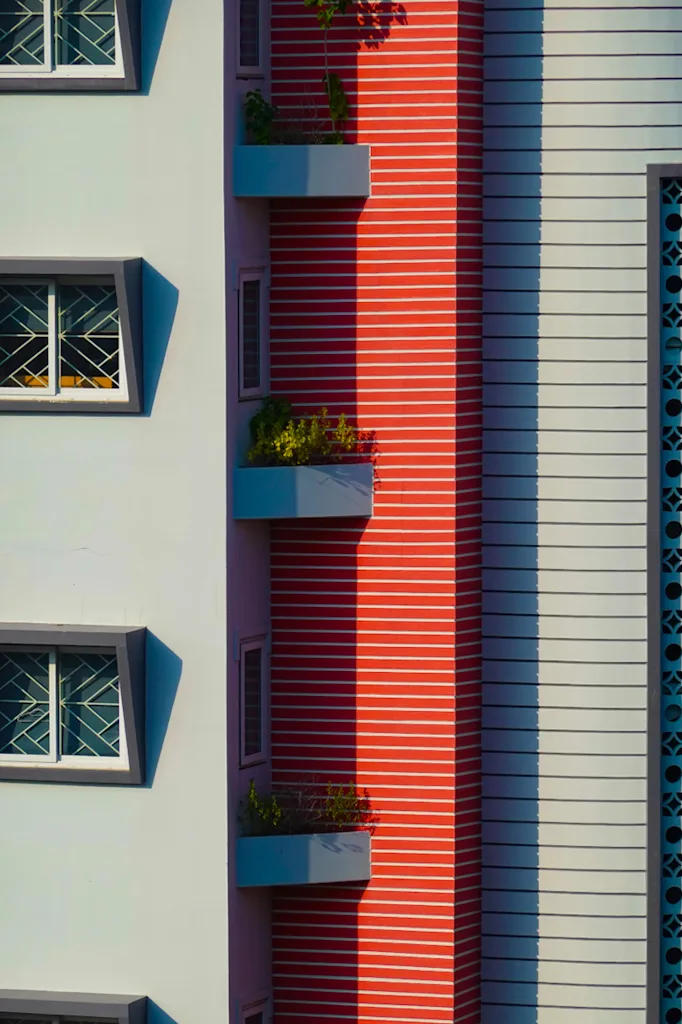 A white facade of angled window hoods beside a column of red horizontal louvres with pale blue planter boxes stepping down it, and a teal breeze block screen at the right edge