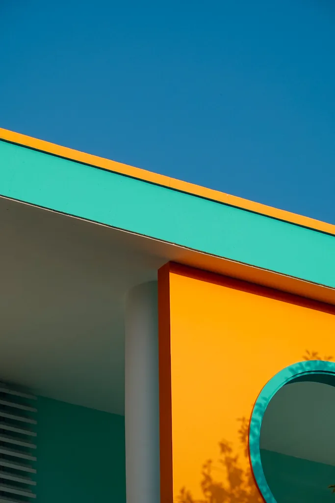 The corner of a building in saturated blocks of colour: an orange fascia, a mint green soffit, and a marigold wall with a teal circular window, against a deep blue sky
