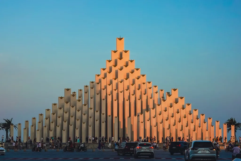 A monument of stepped concrete pillars lit orange by the setting sun, palms at either end, with people on the steps and traffic crossing the foreground