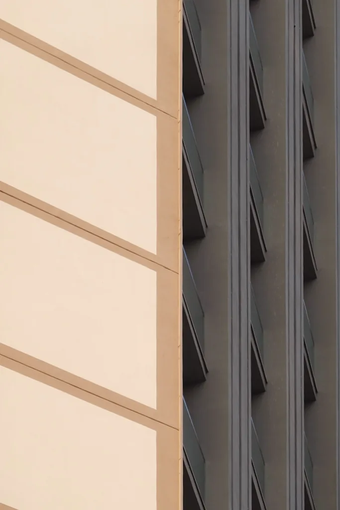 Two facades meeting at a corner: a cream wall with thin horizontal banding at left, and a grey wall of angled window hoods in shadow at right